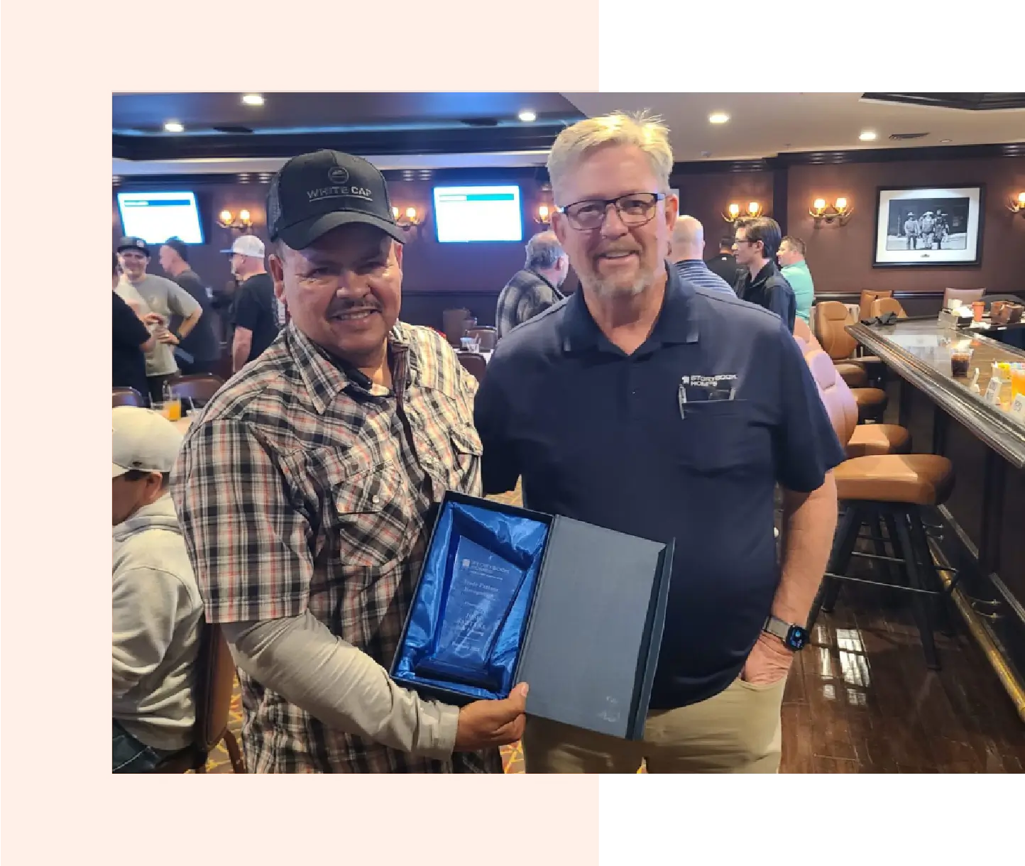 Two men posing with an open award plaque in a lively bar setting.